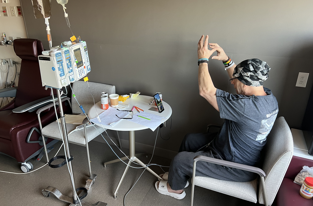 Howard Aaron sits in an armchair in his patient room, facing an iPhone with a video call, raising his arms above his head to instruct a client during a virtual workout. 